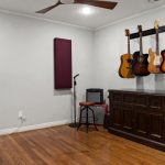 A music room with a wooden floor, a dark wooden cabinet, four guitars hanging on the wall, a microphone stand, a small amplifier, a stool, and a brown leather chair. A ceiling fan and acoustic panel are also visible.