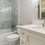 A modern bathroom with a glass-enclosed bathtub and shower, a white toilet, a cream-colored vanity with a marble countertop, a sink, gold fixtures, and a mirrored cabinet above the sink.