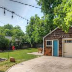 A backyard with a detached garage, string lights overhead, a concrete driveway, green lawn, trees, and a small red play structure in the distance. A flower bed borders the house on the left.