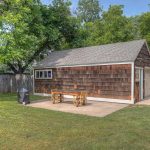 A brown wooden shed with a gray shingled roof stands in a grassy backyard next to a tall wooden fence. There is a picnic table and benches on a concrete patio outside the shed. Trees provide shade in the background.