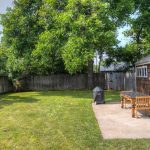 A backyard with a grassy lawn, trees, and a wooden fence. There is a small patio with a wooden table, benches, and a covered grill next to a shingled garage or shed.