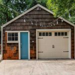 A brown shingle garage with a light blue door, white-framed window, and white garage door. String lights hang overhead, and a wooden picnic table sits on the lawn to the left, surrounded by trees and a wooden fence.