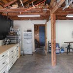 A garage with exposed wooden ceiling beams, white storage cabinets, a workbench, drawers, a metal filing cabinet, a standing fan, boxes, and an exercise bike on a concrete floor.