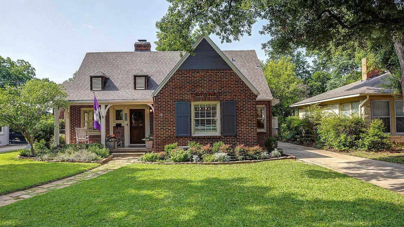 A charming brick house with a gabled roof, front porch, and shuttered windows sits amid a well-kept lawn and landscaped garden, with a driveway on the right and trees and neighboring house in the background.