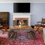 A cozy living room with gray armchairs, leather chairs, a wooden coffee table, a vibrant red patterned rug, a fireplace with a bouquet of flowers on the mantel, a piano, and a large TV mounted above the mantel.