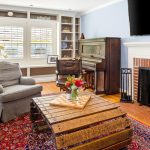A cozy living room with a fireplace, wooden coffee table, gray armchair, upright piano, built-in shelves, large window with shutters, and a colorful rug on a hardwood floor. A vase of flowers sits on the table.