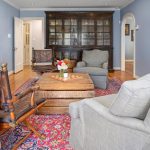 A cozy living room with blue walls, a wooden cabinet, fireplace, two gray armchairs, a wooden rocking chair, a wooden coffee table with flowers, and a red patterned rug on a hardwood floor.