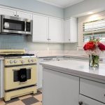A bright kitchen with white cabinets, a vintage yellow oven, a microwave, and a checkered floor. A vase with red and pink flowers sits on a marble-topped island. Sunlight streams through a window with shutters.