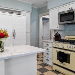 A bright kitchen with white cabinets, a marble countertop island with a vase of flowers, a vintage cream-colored stove, a microwave, and a checkered tile floor. Pale blue walls and a white fridge complete the space.