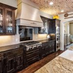 A spacious kitchen features dark wood cabinets, a stainless steel gas stove with a decorative hood, a granite island with a sink, and brick ceiling accents. Wall decor includes vintage wooden paddles.