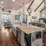 Spacious, elegant kitchen with a granite island, farmhouse sink, pendant lights, brick ceiling, hardwood floor, and open plan leading to a cozy living room with a fireplace and dining area.