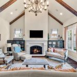 A cozy living room with vaulted ceiling, exposed wood beams, a chandelier, white walls, a fireplace beneath a mounted TV, sofas, armchairs, lamps, side cabinets, and large windows with pink curtains letting in natural light.