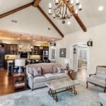 Spacious living room with vaulted ceiling, exposed wooden beams, two sofas, a coffee table, a chandelier, and an open view into a kitchen with barstools and pendant lights.