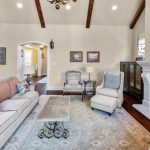 Cozy living room with vaulted ceiling, exposed wood beams, a fireplace with a TV above, beige sofas with floral pillows, an armchair, a coffee table, and large rug covering wooden floors. Light streams in from adjacent rooms.