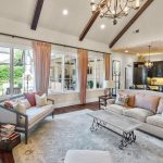 Bright, airy living room with vaulted ceiling, exposed beams, large windows with peach curtains, light sofas with pastel cushions, a coffee table, and open view into the kitchen with pendant lights.