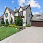 A two-story white brick house with a brown tile roof, wooden garage door, manicured lawn, and colorful flowerbeds under a blue sky with scattered clouds.