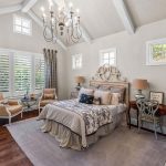 A bright, elegant bedroom with a large bed, two armchairs by a window, a desk with a chair, hardwood floors, a chandelier, and neutral décor in beige and white tones. Natural light fills the room.