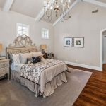A spacious, elegant bedroom with a high, vaulted ceiling, chandelier, queen bed with patterned bedding, and two nightstands. Hardwood floors, a large gray rug, and classic furniture complete the cozy, bright space.