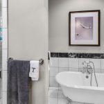 Modern bathroom with a walk-in glass shower, a freestanding white bathtub, a towel hanging on a rail, and a framed artwork above the tub; white tile and marble accents.