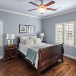 Bright bedroom with light blue walls, a wooden sleigh bed with white bedding, two nightstands with lamps, hardwood floors, wall art, ceiling fan, and windows with white shutters letting in natural light.