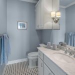 A bathroom with light blue walls, a white toilet, white vanity with marble countertop, silver faucets, a large mirror, wall-mounted lights, and black-and-white checkered floor tiles. Blue towels hang by the shower and sink.