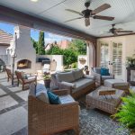 A covered patio with wicker furniture, a coffee table, potted plants, ceiling fans, and a TV. An outdoor fireplace, grill, and additional seating are in the open patio area, surrounded by greenery and white brick walls.