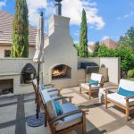 A cozy outdoor patio with wooden furniture, blue and white cushions, a white brick fireplace, built-in grills, potted plants, and a view of a green backyard under a sunny sky.
