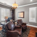 A cozy home office with dark wood furniture, two brown leather chairs, a desk, bookshelves, a fireplace, wall art, and large windows with white shutters. Warm lighting and a patterned rug complete the space.