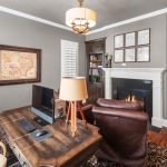 A cozy home office with a wooden desk, two leather chairs, computer, bookshelves, a fireplace, framed maps on the walls, and a warm ambiance from the fire and ceiling light.