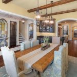 Spacious dining room with a wooden table, upholstered chairs, chandeliers, and exposed ceiling beams. The room opens to a hallway and living area with hardwood floors and arched doorways.