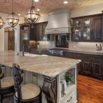 Spacious kitchen with a large granite island, four ornate chairs, dark wooden cabinets, stainless steel appliances, pendant lights, and a brick-patterned ceiling. Decor includes yellow vases and built-in oven and microwave.