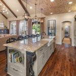 A spacious kitchen and living area with a large island, granite countertops, open shelves, dark wood cabinets, chandeliers, exposed brick ceiling, hardwood floors, and a cozy sitting area with a fireplace and TV.