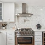 Modern kitchen with white cabinets, a stainless steel oven and range hood, marble tile backsplash, countertop appliances, a wine fridge, and a dining area visible through an arched doorway.