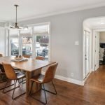 A dining room with a wooden table, six brown leather chairs, a modern chandelier, hardwood floors, large windows, and a hallway leading to a home gym with exercise equipment.