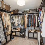 A neatly organized walk-in closet with shelves, drawers, and hanging clothes on both sides. Shoes, bags, and storage baskets are arranged on the lower shelves and top racks. Carpeted floor and a ceiling light are visible.