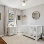 A bright, modern nursery featuring a white crib, a round mirror, a small table with a lamp by the window, framed wall art, a shelf with stuffed animals, and a basket of toys on a soft rug over wooden floors.