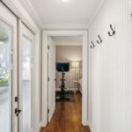 A hallway with wooden floors and white paneled walls, featuring several black coat hooks on the right. Glass double doors on the left overlook a yard. At the end of the hall is a room with a stationary bike and a TV.