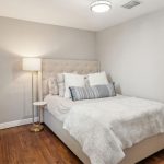 Small bedroom with a neatly made bed featuring white and gray bedding, several pillows, a tufted headboard, a floor lamp, a small round side table, hardwood floors, and light gray walls.