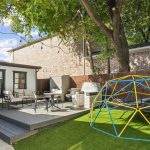 A backyard with artificial grass features a blue and yellow climbing dome, a gray patio deck with outdoor furniture, string lights overhead, a grill, and mature trees providing shade.