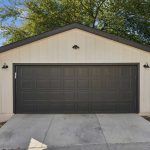 A detached two-car garage with a dark gray door, white siding, and a sloped roof. It is bordered by wooden fences on both sides, with a concrete driveway and trees visible in the background.