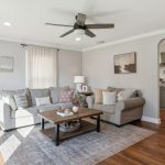 A bright living room with a gray sectional sofa, beige armchair, wooden coffee table, light rug, floor lamp, wall art, and hardwood floors. An open archway leads to a modern kitchen in the background.