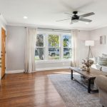 A bright living room with wooden floors, a large window with curtains, a gray sectional sofa with cushions, a coffee table, a floor lamp, wall art, ceiling fan, and a TV on a cabinet with decorative plants.
