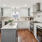 Modern kitchen with white cabinets, stainless steel appliances, a large island with a plant, marble backsplash, wood floors, and pendant lighting over the sink area.