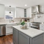 Modern kitchen with white cabinets, stainless steel appliances, a farmhouse sink, a wine fridge, an island with a plant, marble backsplash, wood floors, and a window letting in natural light.
