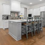 Modern kitchen with white cabinets, stainless steel appliances, a gray island with three barstools, light wood floors, and a decorative tray with lemons on the counter. A hallway and dark front door are visible in the background.