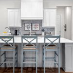 Modern kitchen with a large white island, three gray bar stools, white cabinetry, stainless steel appliances, and a light wood floor. Two framed pictures and a coffee maker are on the counter.
