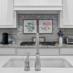 A modern white kitchen with a sink and faucet in the foreground, a granite countertop, white cabinets, a toaster oven, coffee maker, and two floral art prints on a gray tile backsplash.