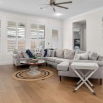 A modern living room with a gray sectional sofa, round coffee table on a woven rug, wall art with family photos, wood flooring, ceiling fan, and large windows with blinds letting in natural light.