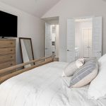 A neatly made bed with white bedding and gray accent pillows sits in a bright bedroom. A wooden dresser with a TV on top and a large mirror are against the wall. Double doors lead to another room.