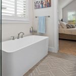 Modern bathroom with a freestanding white bathtub, glass shower, towel rack, wall art, and a beige geometric rug. An open door reveals a bedroom with a bed and windows with plantation shutters.